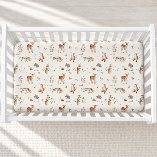 A fitted crib sheet with a woodland animal print is shown inside a white wooden crib, viewed from above. The print features brown deer, foxes, rabbits, hedgehogs, and squirrels with green leaves on an off-white background.