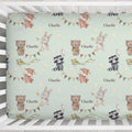 A crib mattress or mattress protector with a whimsical woodland pattern featuring teddy bears, rabbits, raccoons, bunting, stars, and the name 'Charlie' repeated, shown inside a white wooden crib.
