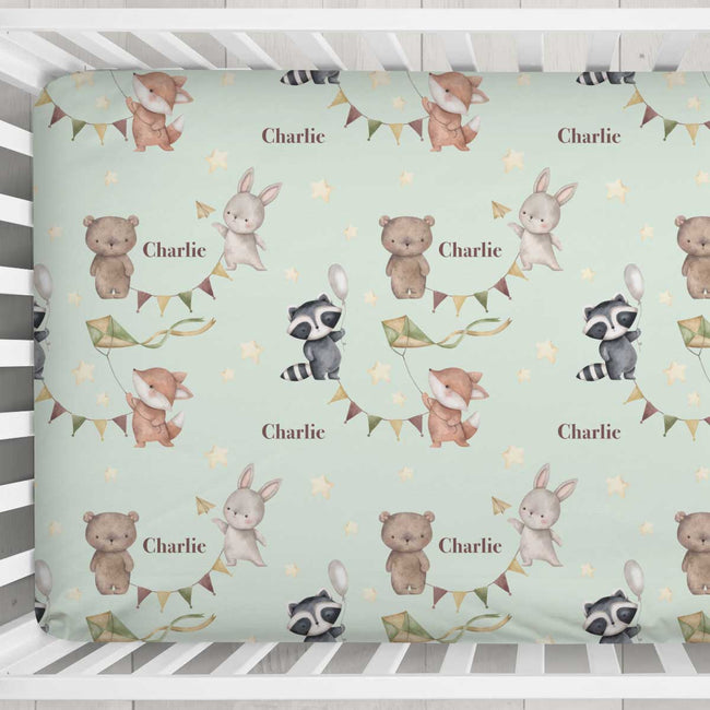 A crib mattress or mattress protector with a whimsical woodland pattern featuring teddy bears, rabbits, raccoons, bunting, stars, and the name 'Charlie' repeated, shown inside a white wooden crib.