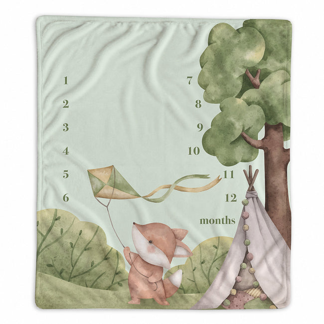 A square baby milestone blanket featuring a watercolor illustration of a fox holding a kite next to a tree and a teepee, with printed numbers 1 through 12 and the word 'months' on a pale mint background.