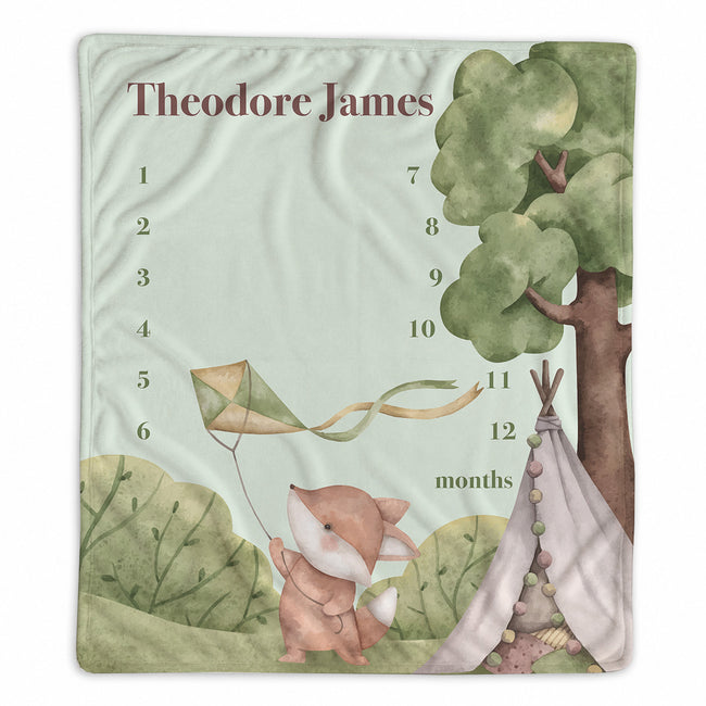 A personalized baby milestone blanket featuring a watercolor woodland scene with a fox, tree, and teepee, alongside the name 'Theodore James' and a numbered months grid from 1 to 12.