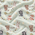 A close-up of a soft fleece blanket with a watercolor woodland print featuring teddy bears, a rabbit, a raccoon, a fox, stars, bunting, and the name 'Charlie'.