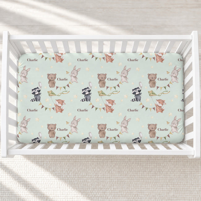 A top-down view of a white wooden crib with a custom printed mattress protector featuring woodland animal illustrations and the name 'Charlie' on a pale mint green background.