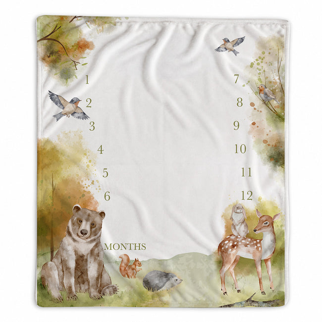 A square baby milestone blanket with a watercolor woodland design, featuring a bear, deer, squirrel, hedgehog, birds, and a clock face showing 12 months.