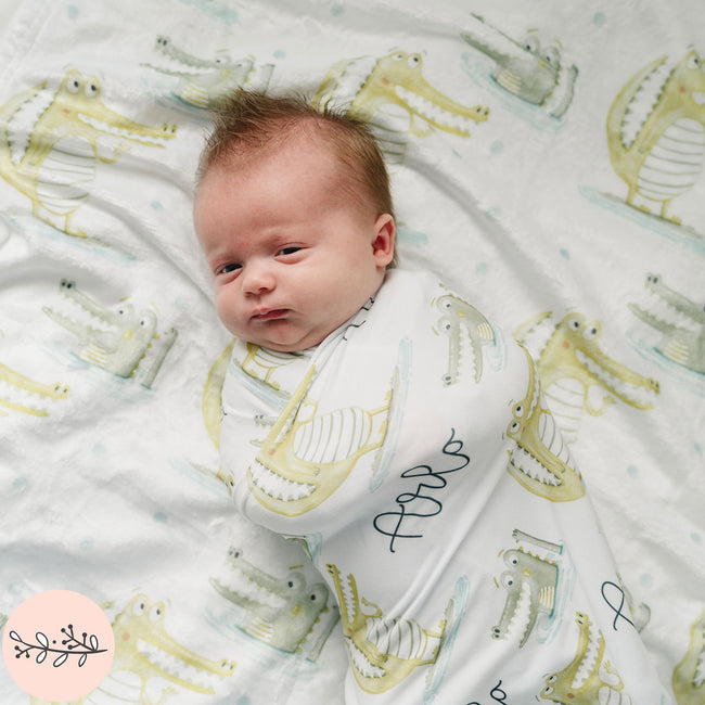 a baby wrapped in a personalized baby swaddle with a cute gator pattern laying on a matching blanket