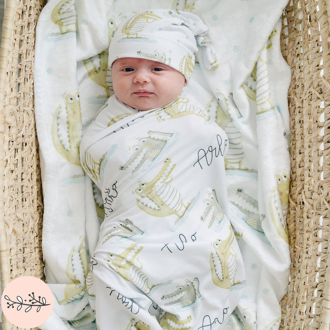 A baby wrapped in a personalized swaddle blanket with alligator print, lying in a basket.