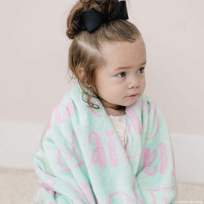 A young child sitting on the floor with a personalized name blanket in peach and bright mint colors
