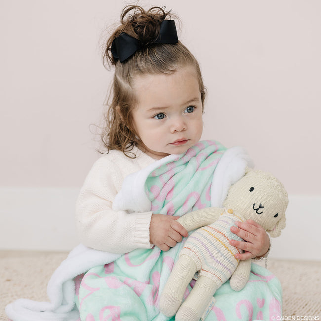 A young child sitting on the floor with a personalized name blanket in peach and bright mint colors, holding a plush toy.