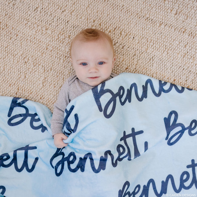 A baby lying on a personalized blue blanket with the name 'Bennett' printed in a cursive script.