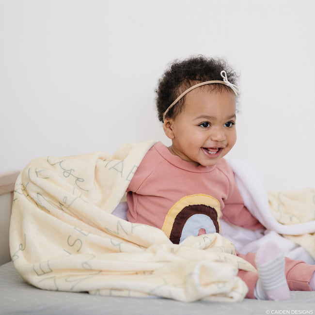 A little girl smiling and wrapped in a soft, plush personalized baby blanket with names