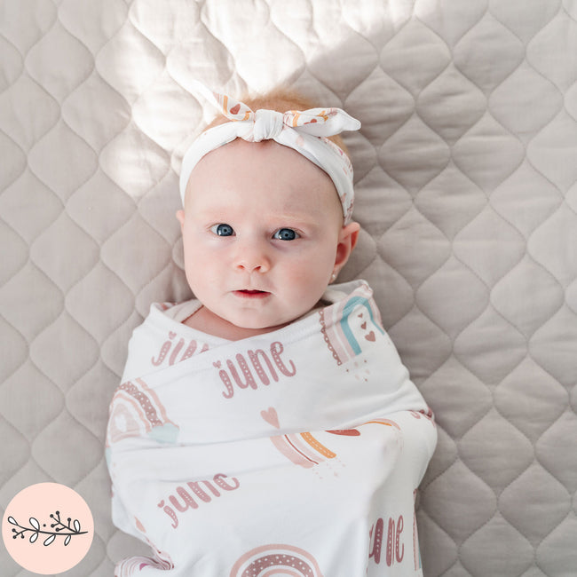 A baby wrapped in a personalized baby swaddle with a boho rainbow design and matching headband.