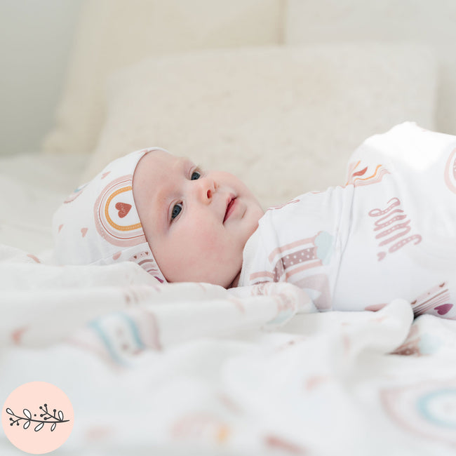 A baby wrapped in a personalized baby swaddle with a boho rainbow design laying on a matching fleece blanket