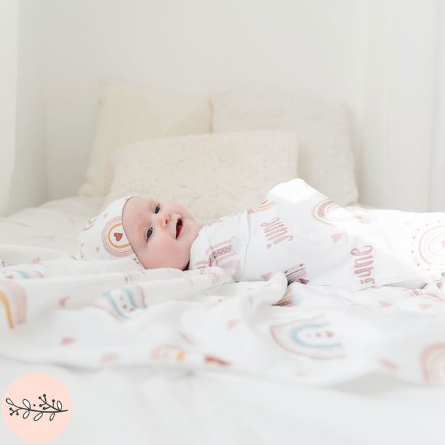 A baby wrapped in a personalized baby swaddle with a boho rainbow design laying on a matching fleece blanket