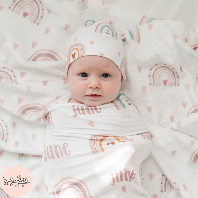 A baby swaddled in a beige blanket with a boho rainbow pattern, lying on a similar blanket with the name 'June' printed on it.