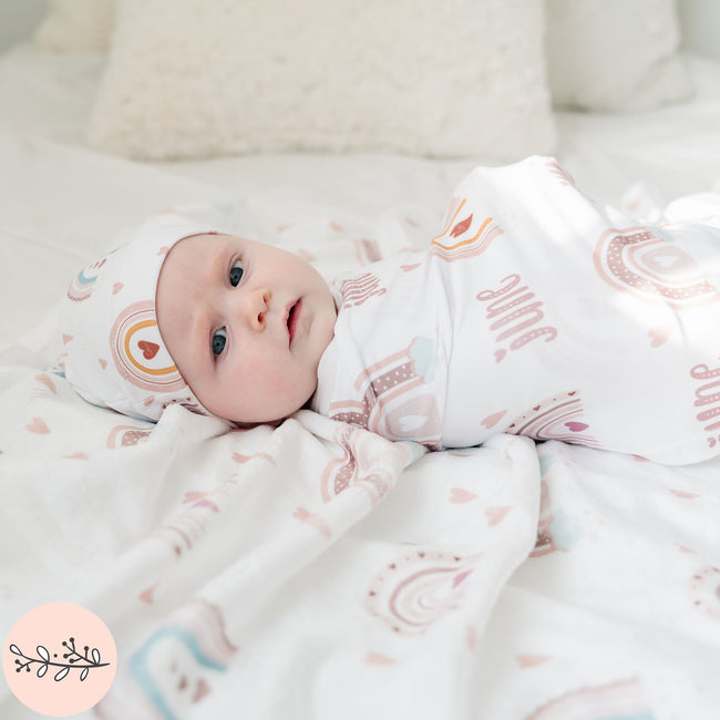 A baby wrapped in a personalized baby swaddle with a boho rainbow design laying on a matching fleece blanket