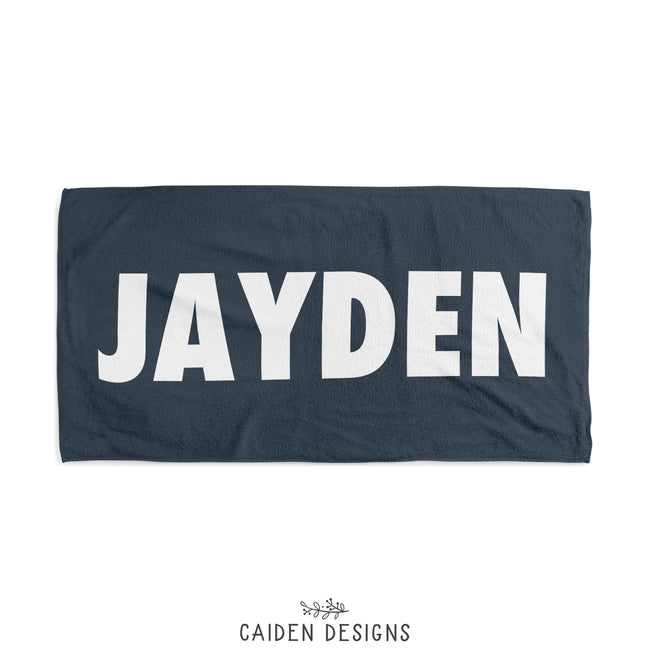 A personalized beach towel with the name 'JAYDEN' printed in large white letters on a dark blue background.