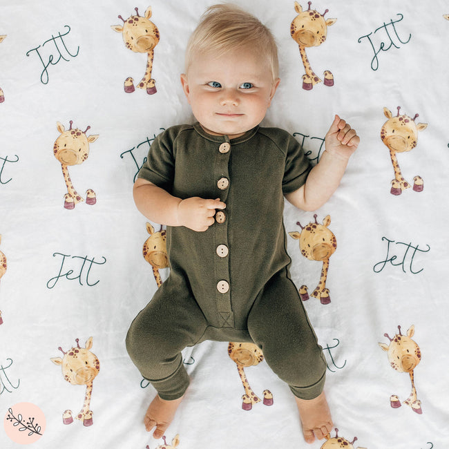A baby lying on a personalized giraffe blanket with the name 'Jett' printed alongside giraffe illustrations.