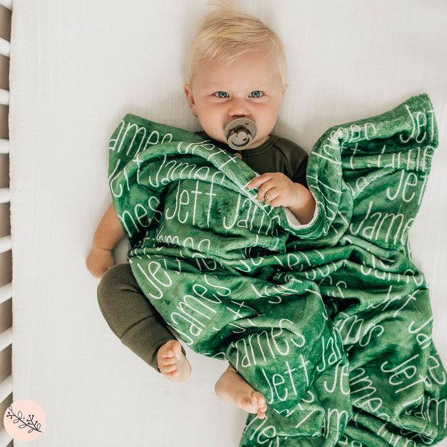 A baby lying on a dark green personalized blanket with the name 'Jett' printed in a white script.