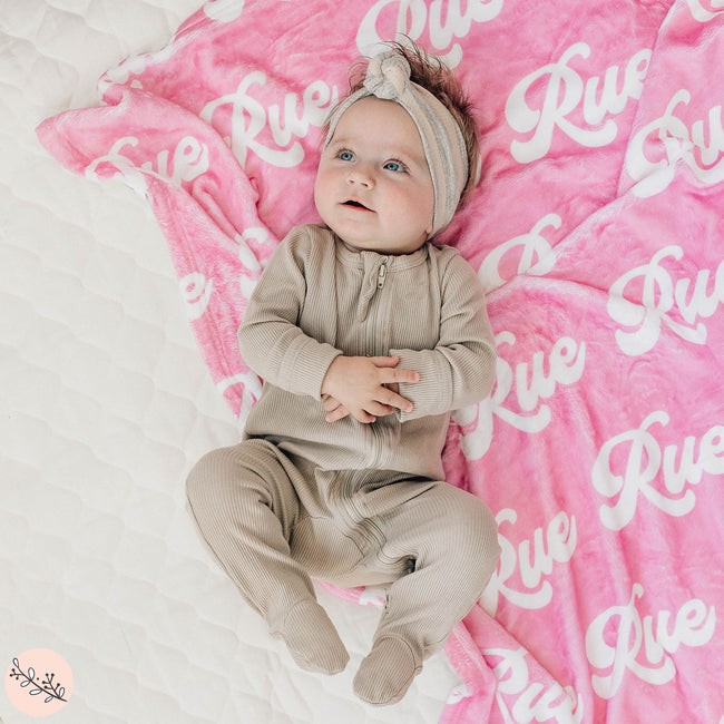 A baby lying on a pink personalized blanket with the name 'Rue' printed in white text.