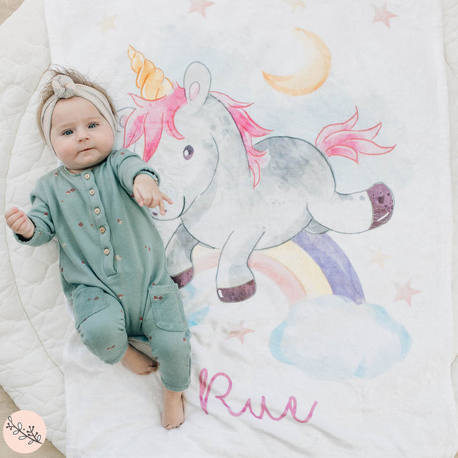 A baby lying on a personalized unicorn blanket with the name 'Rue' printed on it, featuring a colorful unicorn design and other playful elements like stars and a rainbow.