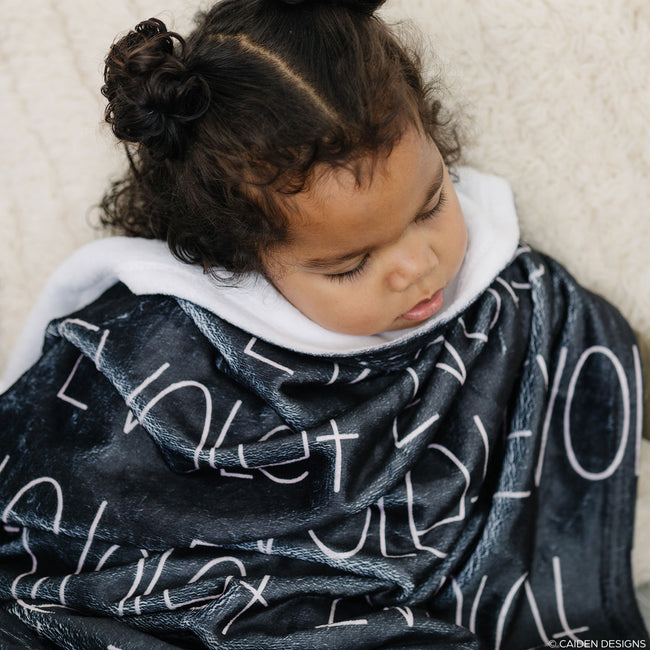 A child wrapped in a black personalized blanket with a blush name printed on it.