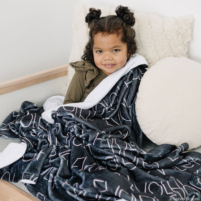 A child wrapped in a black personalized blanket with a blush name printed on it.