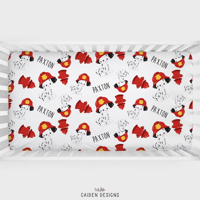A crib sheet with a pattern of dalmatian dogs in firefighter uniforms, with the name 'Paxton' printed on it. The design is mainly in red and white, with a white crib background.