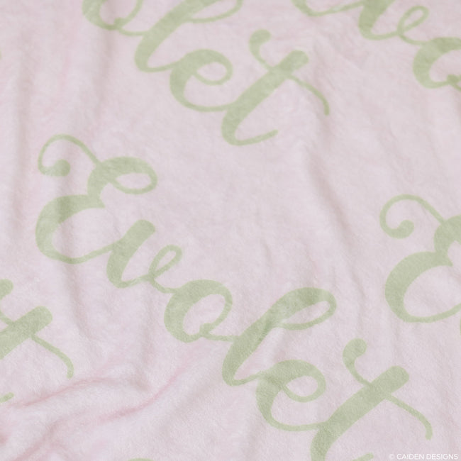 A closeup of a soft pink personalized baby blanket with a light green repeated name pattern 