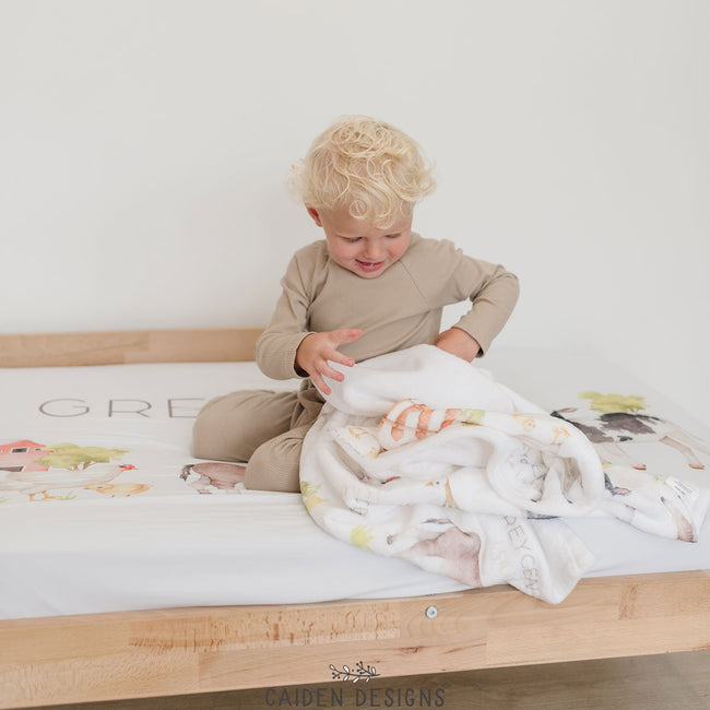 A little boy on a personalized toddler bed sheet and a matching personalized baby blanket with farm animal designs, including a sheep, cow, and pig, along with the name 'Greycen'  printed on it.