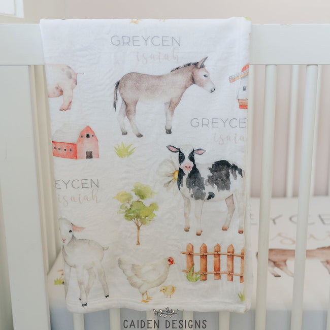 A personalized baby blanket with farm animal designs, including a sheep, cow, and pig, along with the name 'Greycen'  printed on it draped over a crib rail