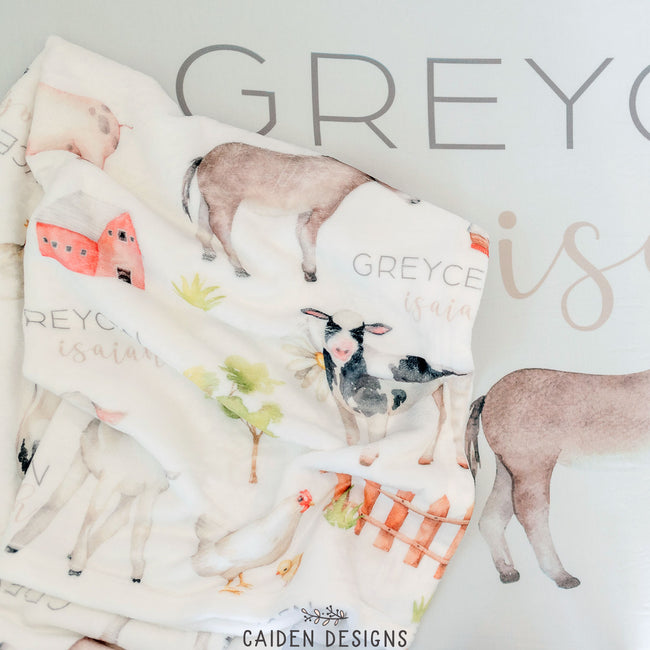 A personalized baby blanket with farm animal designs, including a sheep, cow, and pig, along with the name 'Greycen'  printed on it.