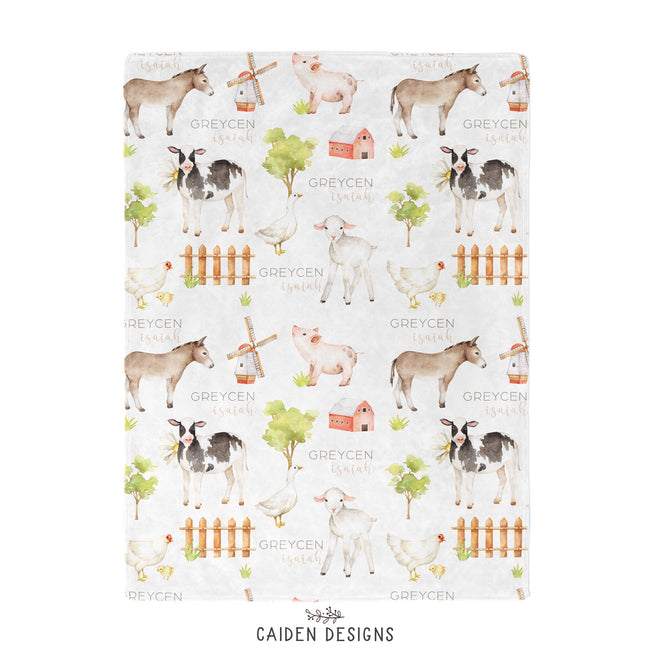 A personalized baby blanket with farm animal designs, including a sheep, cow, and pig, along with the name 'Greycen'  printed on it.