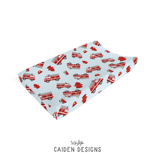 A changing pad cover with a light blue background, printed with red fire trucks, firefighters, helmets.