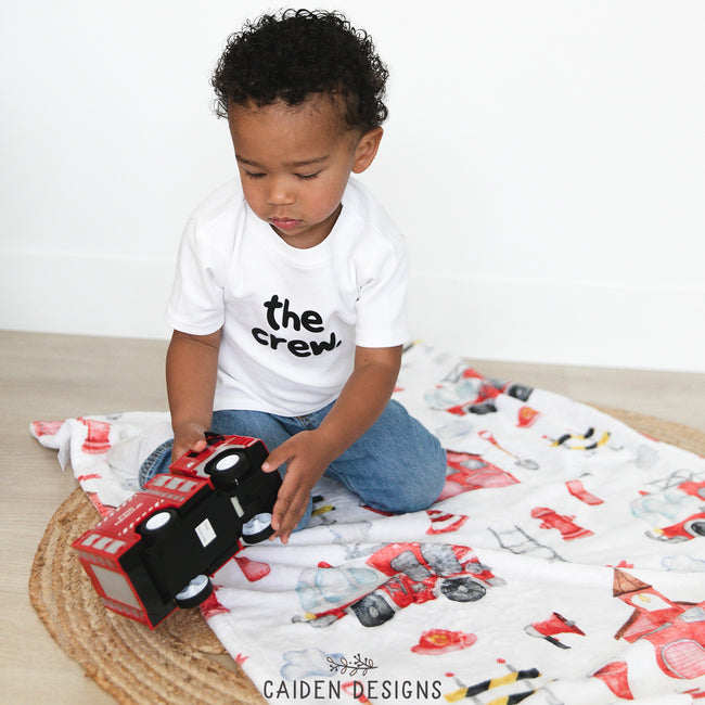 A young child playing with a toy on a personalized blanket featuring firefighting themes and characters.
