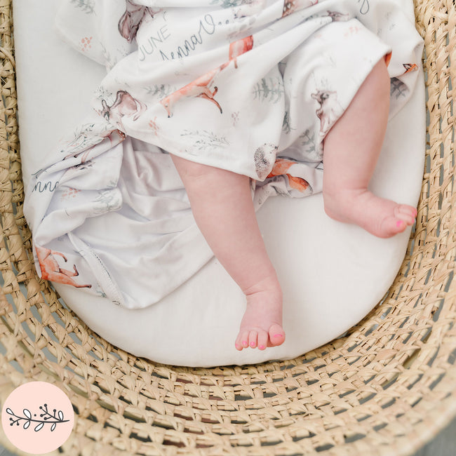 A baby with a blanket featuring woodland animal prints, lying in a wicker basket.