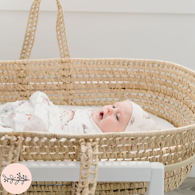 A baby swaddled in a blanket with woodland animal prints, lying in a wicker basket.