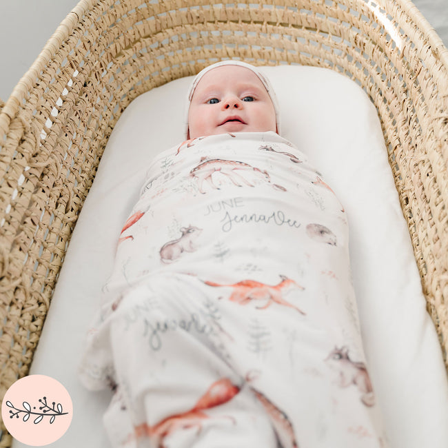 A baby swaddled in a blanket with woodland animal prints, lying in a wicker basket.