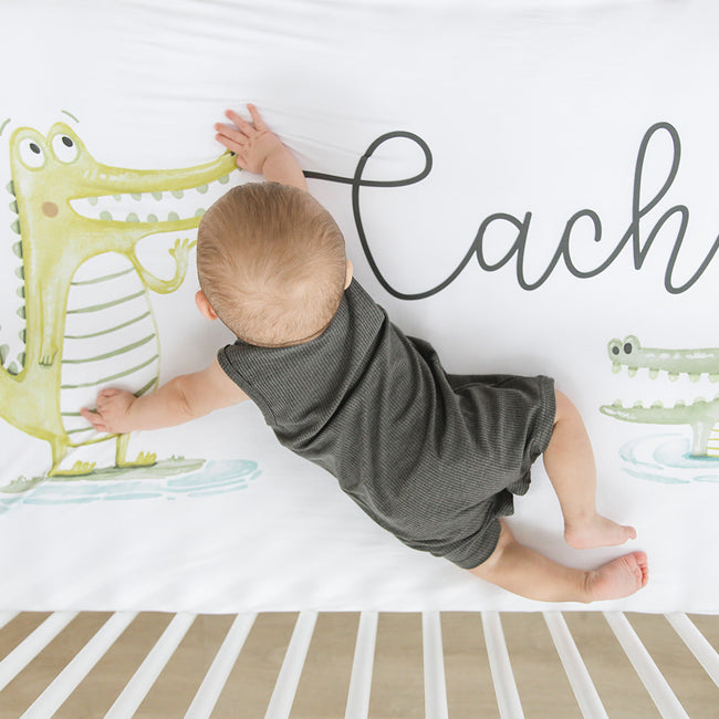 A baby lying on a personalized crib sheet with a printed alligator character.