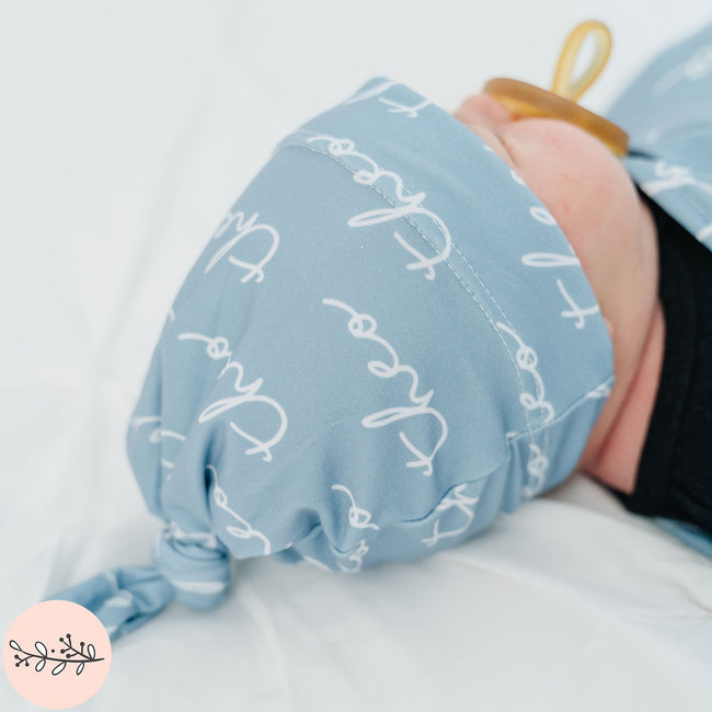 A baby wearing a blue personalized baby hat with the name Theo in a handwritten font
