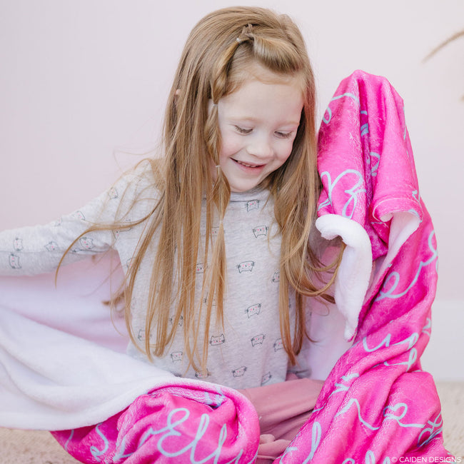 A young girl smiling and wrapped in a pink personalized blanket with a heart interlaced with the aqua names