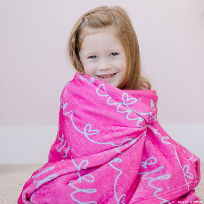 A young girl smiling and wrapped in a pink personalized blanket with a heart interlaced with the aqua names