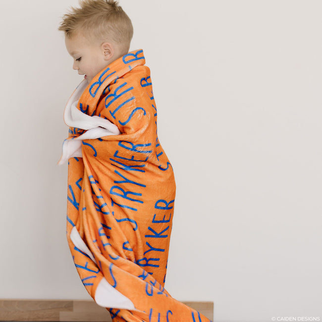 A child wrapped in an orange personalized blanket with blue text standing against a white wall.
