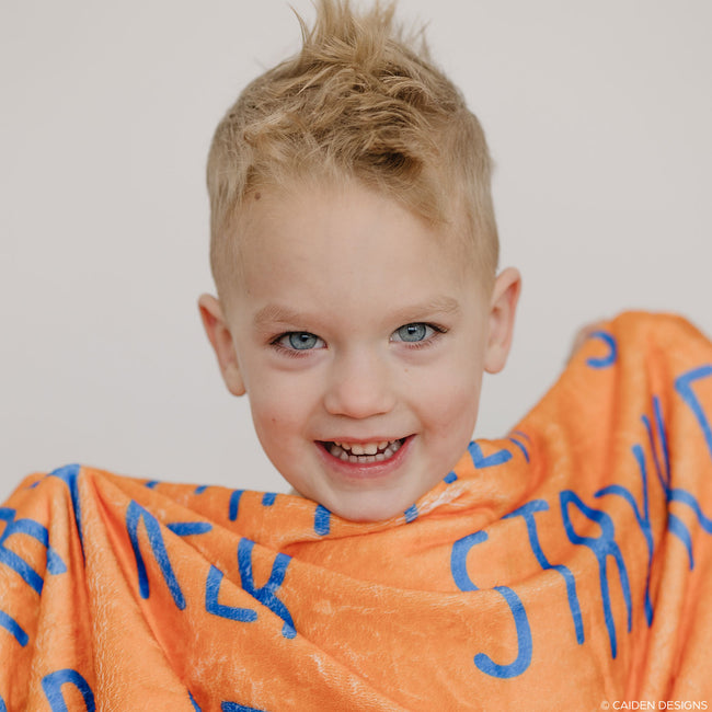 A boy with an orange personalized blanket with cobalt blue letters