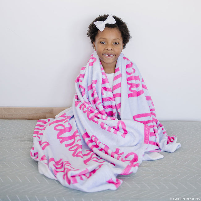 A child sitting on a bed wrapped in a personalized blanket with hot pink names on it.