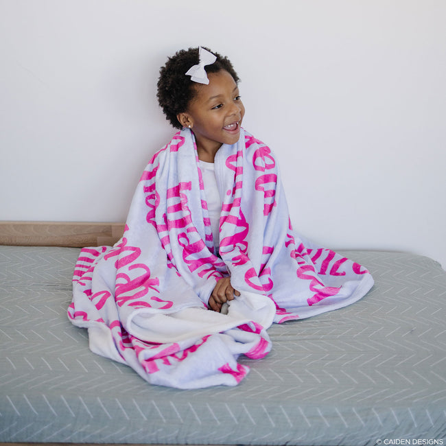 A child sitting on a bed wrapped in a personalized blanket with hot pink names on it.