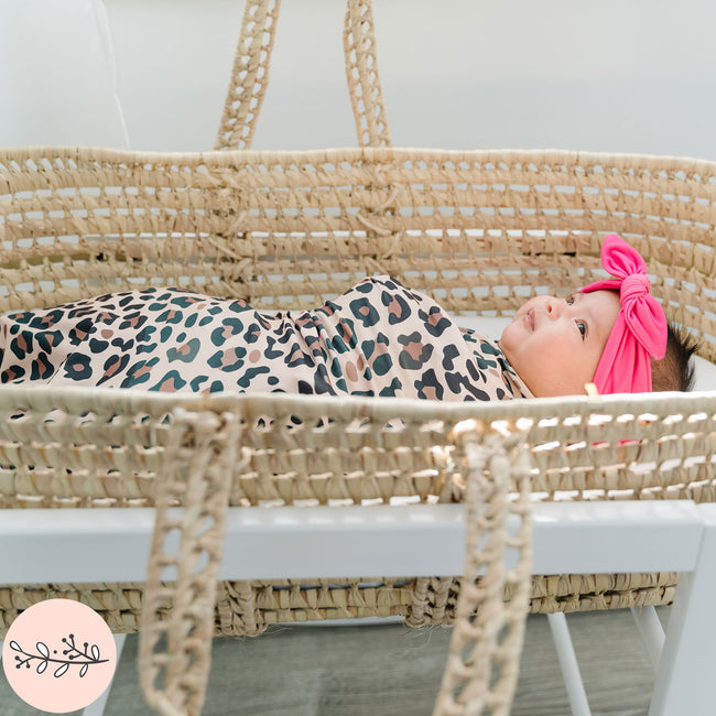 A baby swaddle blanket with brown leopard print design, displayed in a woven basket, with a baby lying inside.
