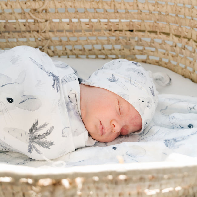 A close up of a newborn baby is swaddled in a gray and white watercolor bunny blanket, lying inside a wicker basket.