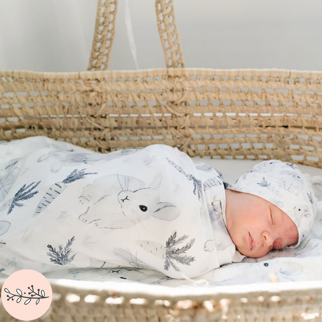 A newborn baby is swaddled in a gray and white watercolor bunny blanket, lying inside a wicker basket.