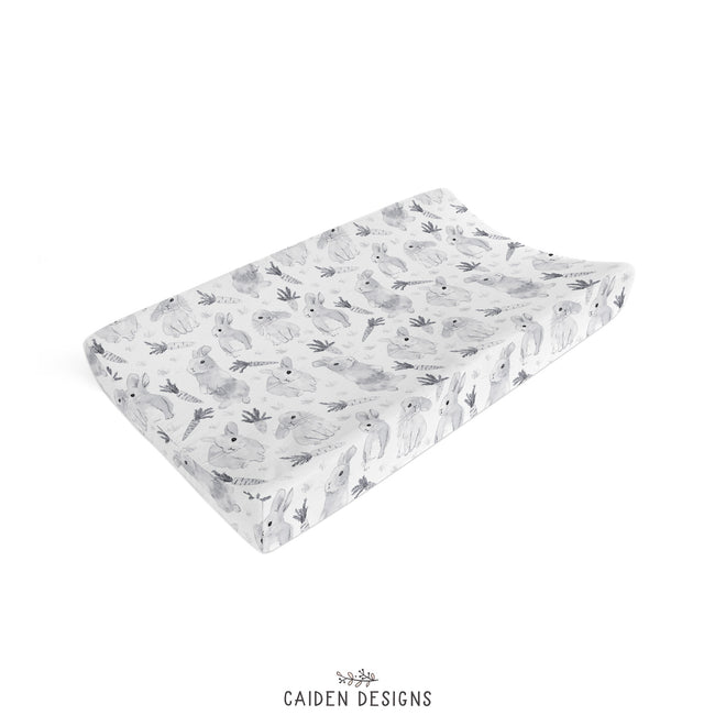 A close-up of baby changing pad cover featuring a repeating pattern of gray watercolor-style rabbits and carrots on a white background. 