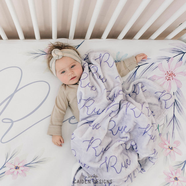 Baby girl lying on a crib with a soft lavender personalized baby blanket with the name Rue on it